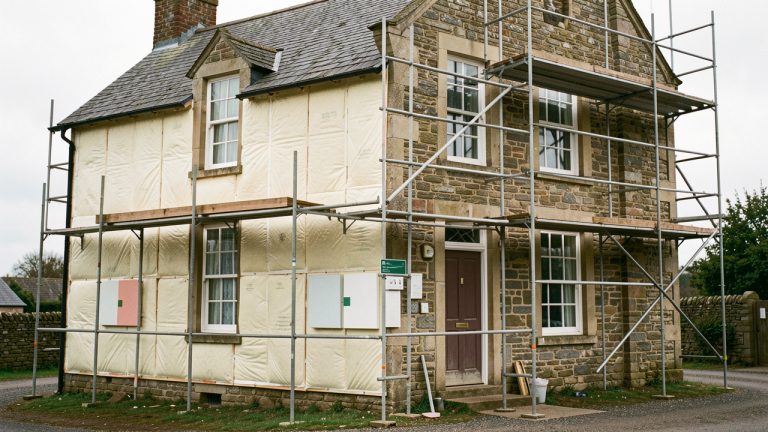 Maison ancienne en rénovation énergétique, avec une façade partiellement isolée, un échafaudage et des panneaux climatiques vierges dans une ambiance documentaire réaliste.